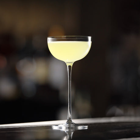 A coupe glass from the Lehmann Glass La Creative London Cocktail Coupe 200ml set evokes elegance as it cradles a pale yellow cocktail, resting on a polished bar surface against a softly blurred background.