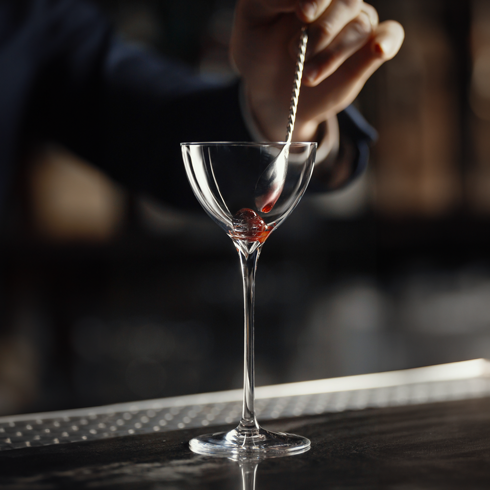 A person uses a twisted mixing spoon to place a maraschino cherry into a Lehmann Glass La Creative New York Nick & Nora Cocktail Glass 170ml.