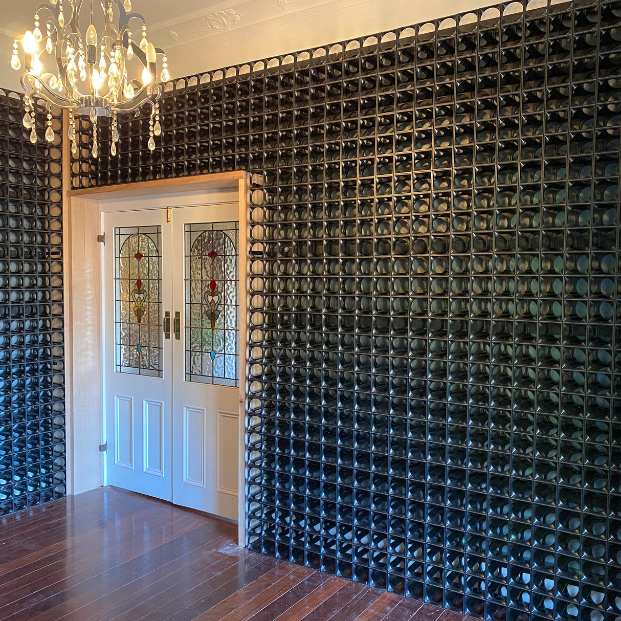 A Stakrax 50 Bottle Plastic Wine Rack - Black by Stakrax, filled with wine bottles, stands by stained glass double doors beneath a chandelier in a room with polished wooden floors.