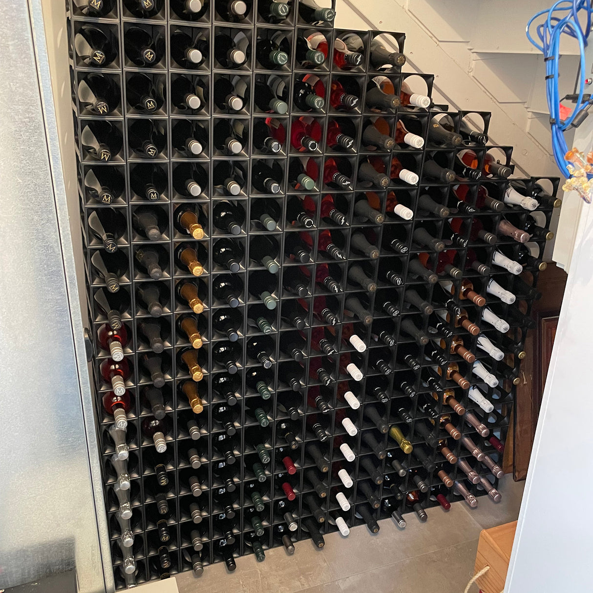 The Stakrax 50 Bottle Plastic Wine Rack by Stakrax, seen under a staircase, organizes numerous wine bottles in horizontal rows and columns. Blue cables are visible hanging on the right side of the image.