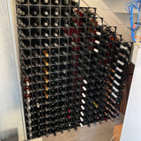 The Stakrax 50 Bottle Plastic Wine Rack by Stakrax, seen under a staircase, organizes numerous wine bottles in horizontal rows and columns. Blue cables are visible hanging on the right side of the image.
