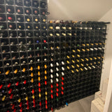 Wine bottles are stored horizontally in a large, black Stakrax 50 Bottle Plastic Wine Rack against two walls of the wine cellar, displaying assorted bottles and labels, with boxes stacked in the corner on the floor.