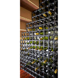 A Stakrax 12 Bottle Modular Plastic Wine Rack in black, featuring multiple wine bottles arranged horizontally in individual square compartments, is set against a wall with wooden shelves above and a wooden floor below.