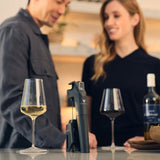 A man and woman stand in a kitchen near two glasses of wine, a wine bottle, and the Coravin Timeless Three+ Wine Preservation System by Coravin (in black) on the counter. Both are smiling and appear to be talking casually.