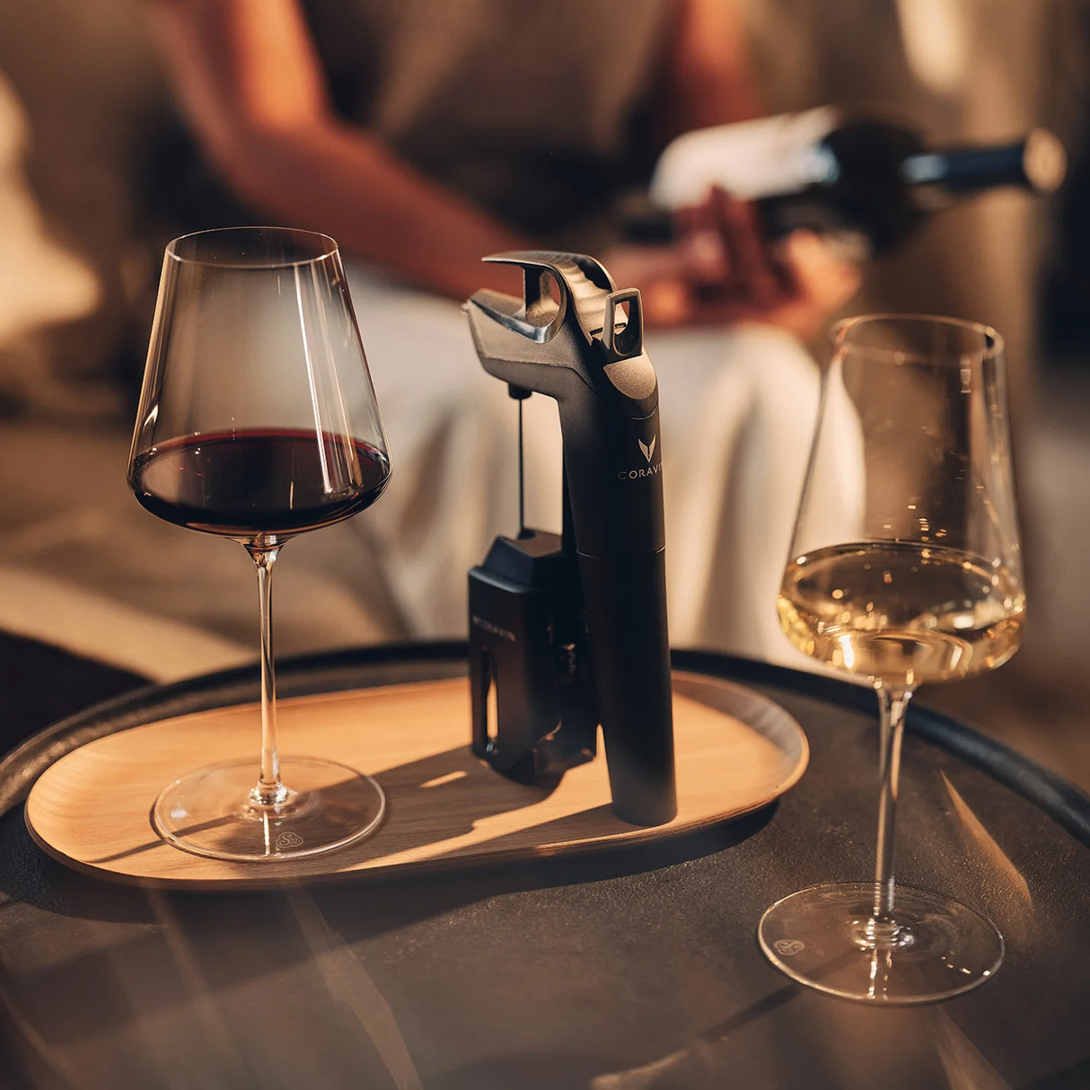 A tray holds two wine glasses—one with red wine, one with white—beside a Coravin Timeless Three+ wine opener, while a person in the background holds a wine bottle. Warm, soft lighting creates a cozy atmosphere, highlighting the sophisticated touch of this advanced wine preservation system.
