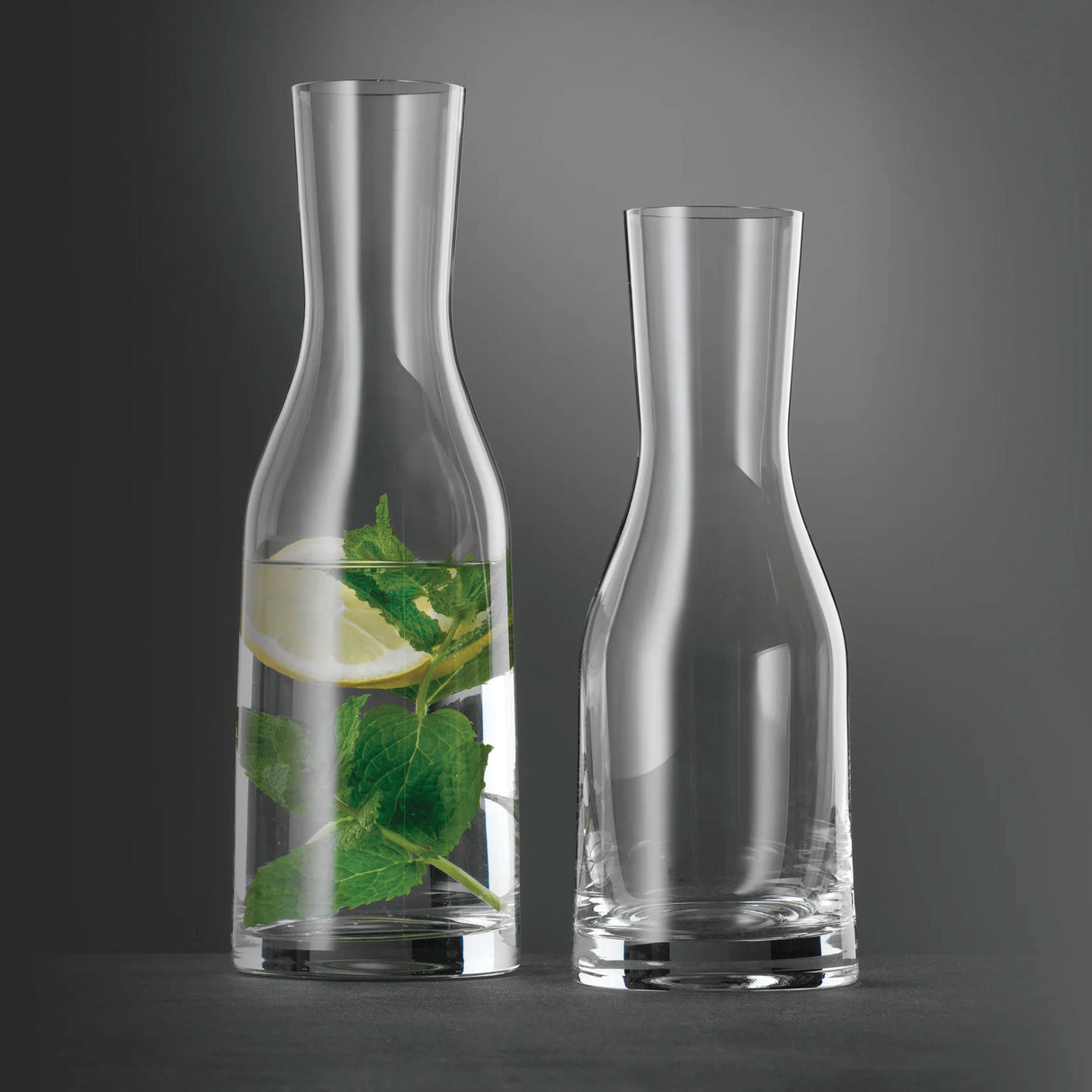 Two clear crystal glass carafes from Spiegelau—the Ultra Wine / Water Carafe 1000ml—are placed side by side; one stands empty, while the other is filled with water, fresh mint leaves, and a slice of lemon. Showcasing the minimalist design of Spiegelau, these elegant carafes stand out beautifully against the gray background, creating a refined and modern scene.